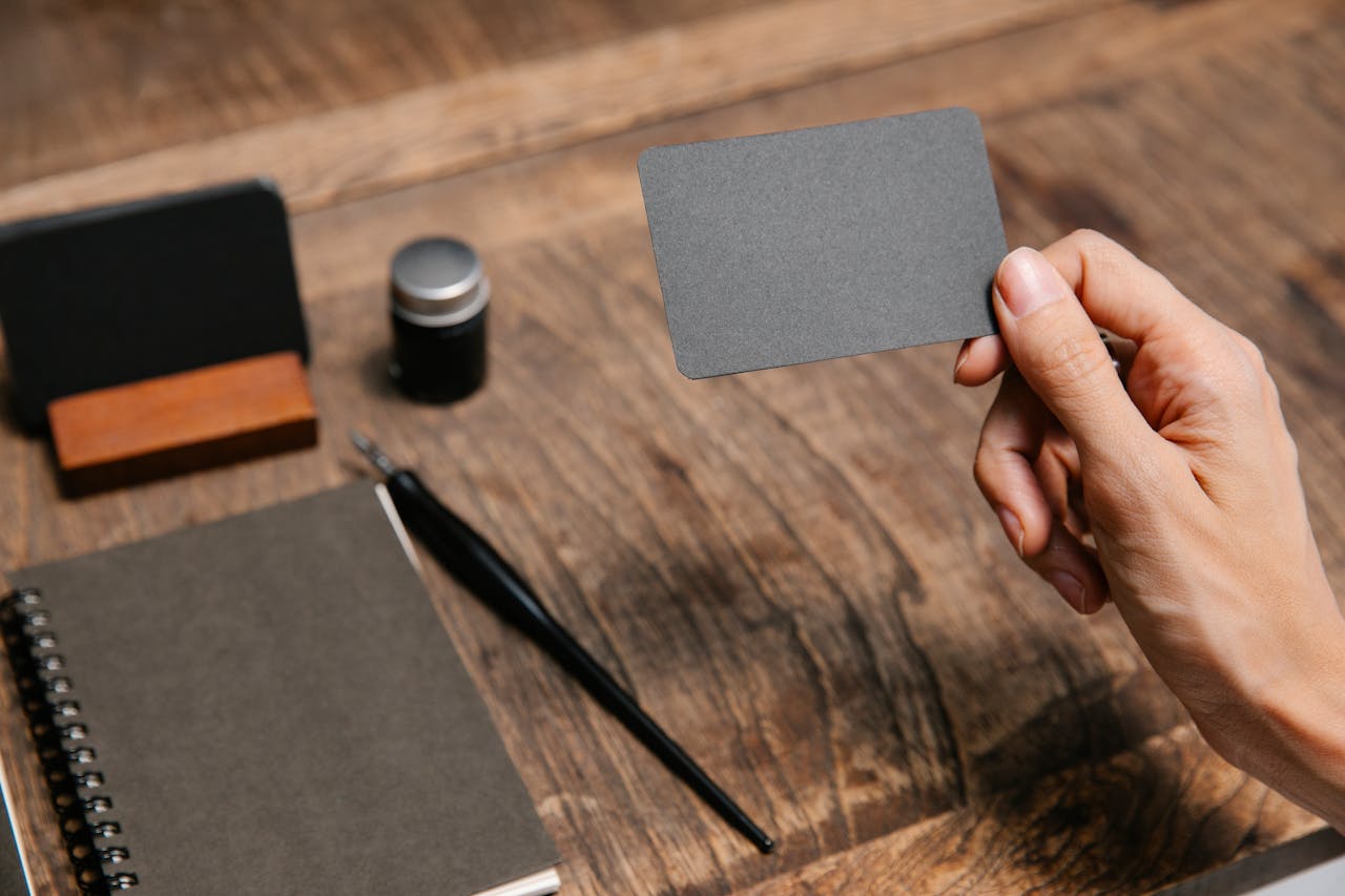A close-up of a hand holding a blank business card above a wooden desk with office accessories.